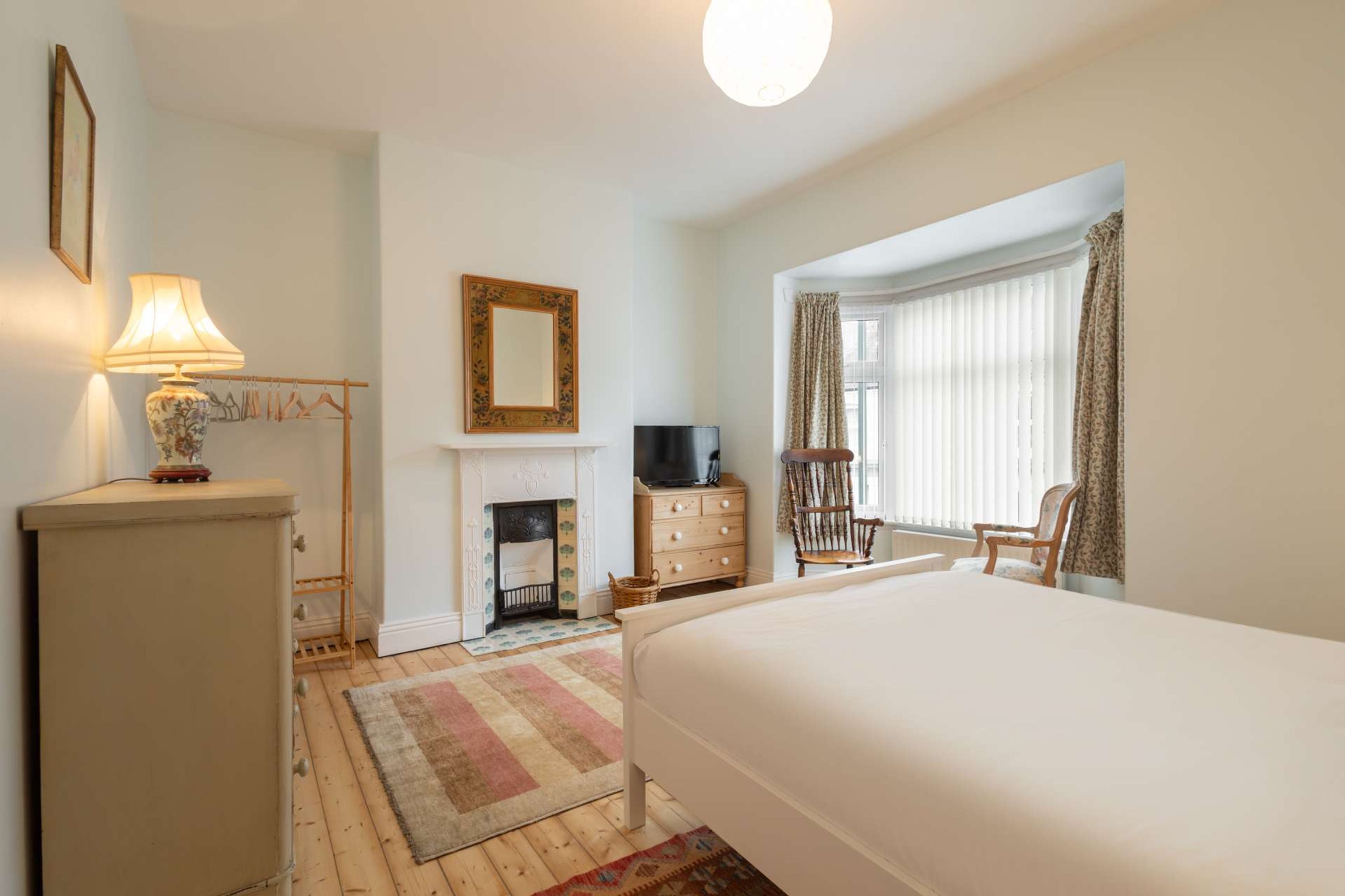 A bedroom with a bed, a dresser, a lamp, a television, a chair, a fireplace, and a window with curtains.