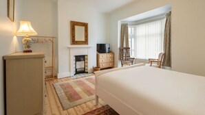 A bedroom with a bed, a dresser, a lamp, a television, a chair, a fireplace, and a window with curtains. - Host Stay Heather House (Saltburn-by-the-Sea)