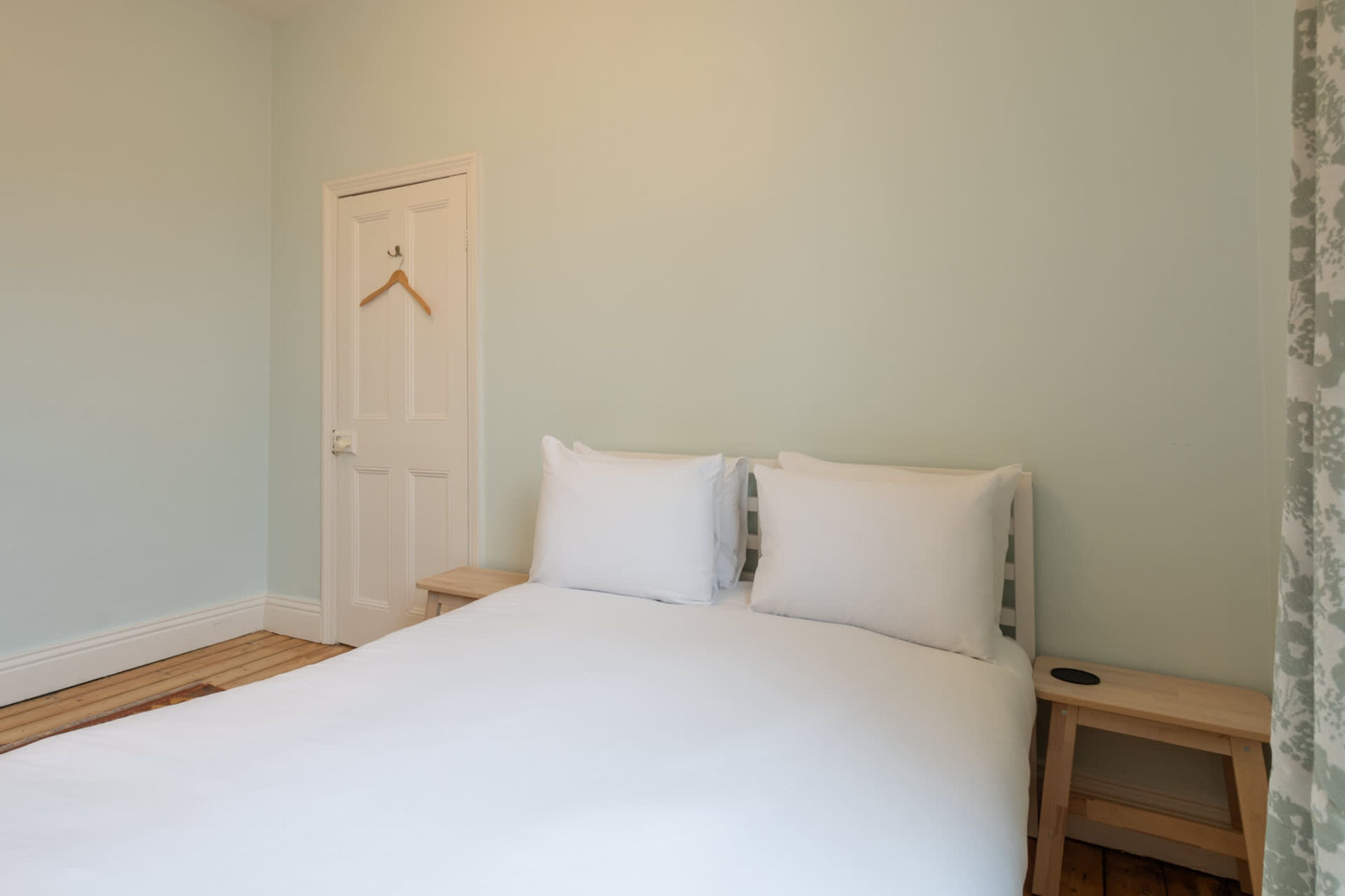 Une chambre à coucher avec un lit, une table de chevet en bois, une porte blanche et un porte-manteau en bois fixé au mur.