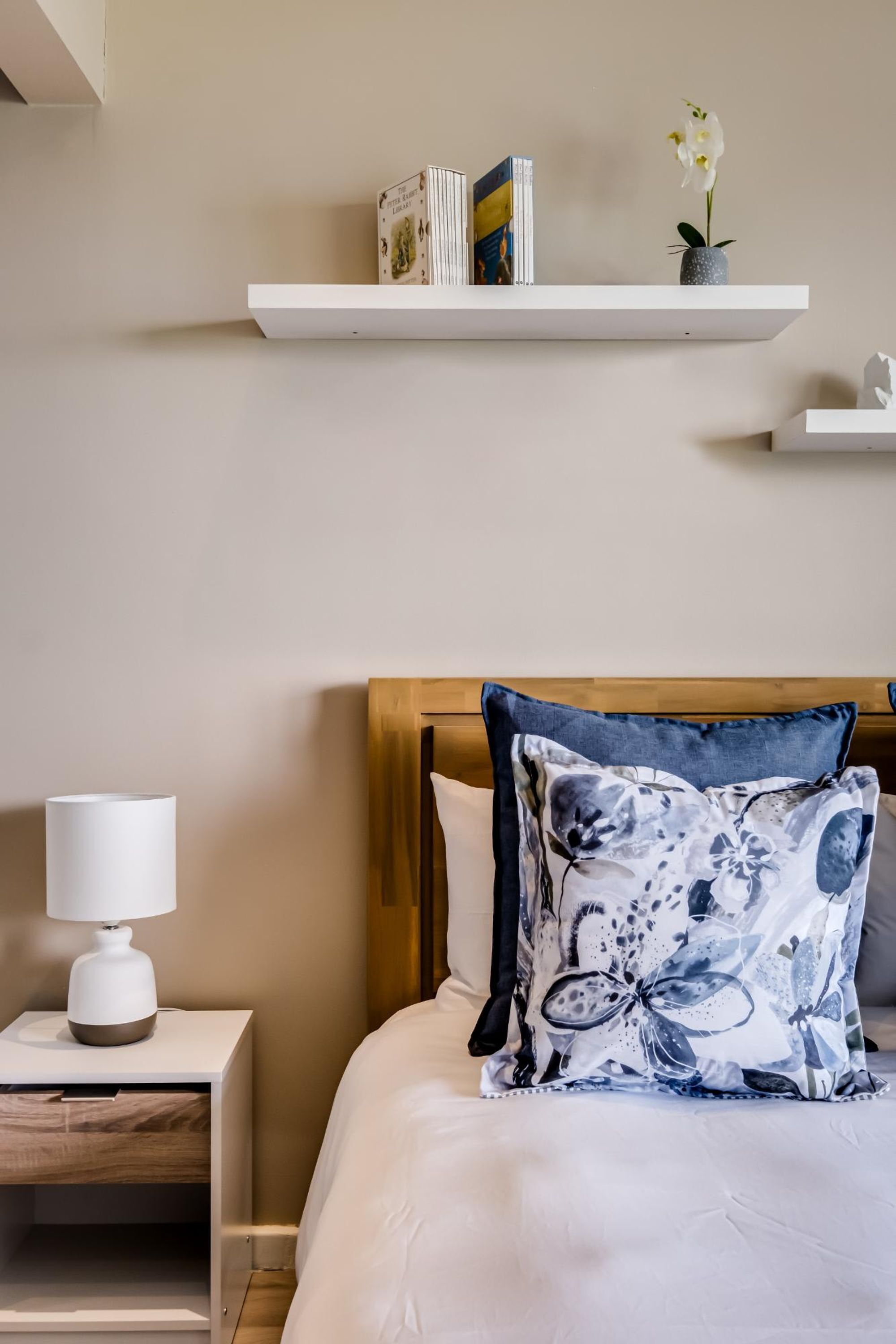 A neatly arranged bedroom with a bed, a bedside table with a lamp, a shelf with books and flowers, and a wooden headboard.