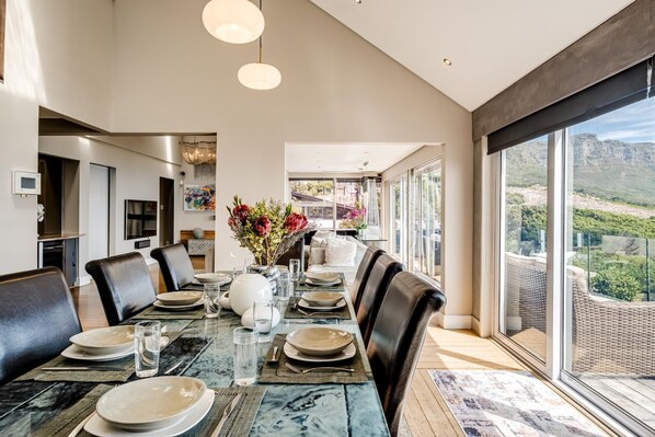 A modern dining area with a long table set for six, surrounded by leather chairs, with a large window offering a scenic mountain view.
