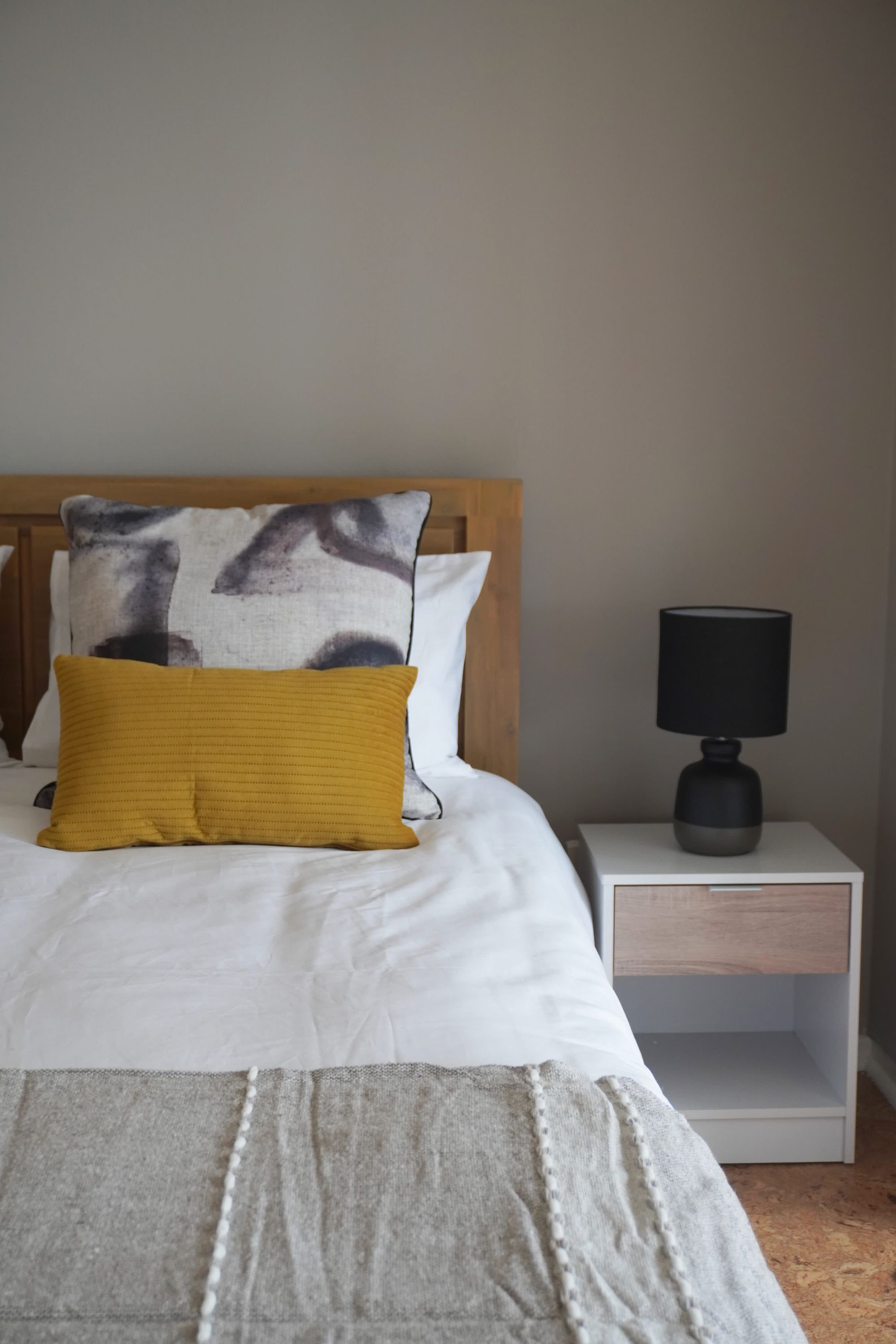 A neatly made bed with white and mustard pillows, a wooden headboard, and a bedside table with a black lamp.