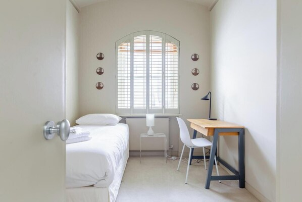Ein kleines, helles Schlafzimmer mit Bett, Schreibtisch, Stuhl und einem Fenster mit Jalousien.