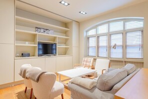 A living room with a sofa, chairs, a coffee table, a TV on a shelf, and a window with blinds.