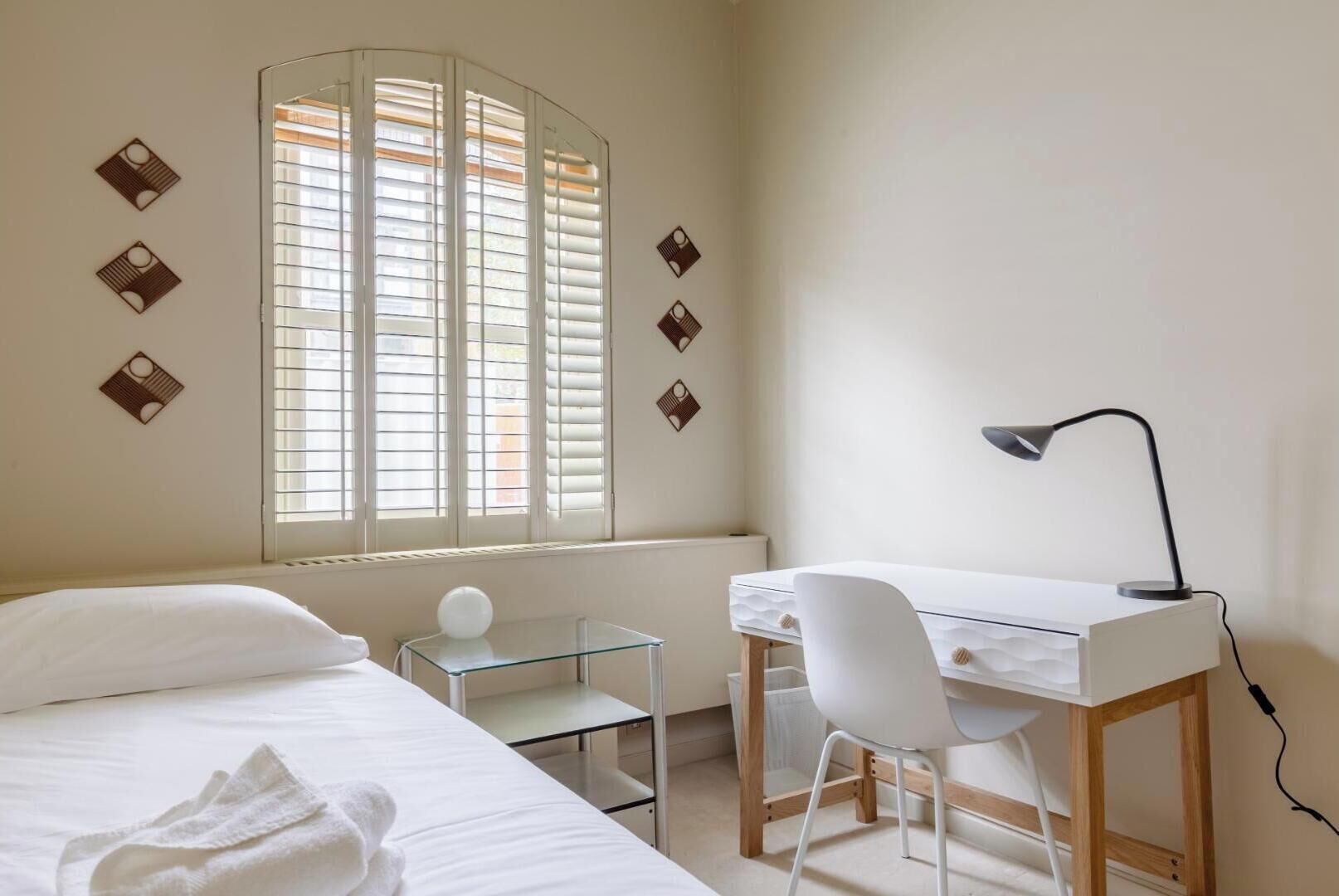 Een slaapkamer met een bed, een bureau, een stoel, een lamp en een raam met jaloezieën.