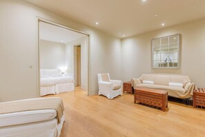 A modern hotel room with a bed, two white armchairs, a sofa, a wooden coffee table, and a large mirror on the wall.