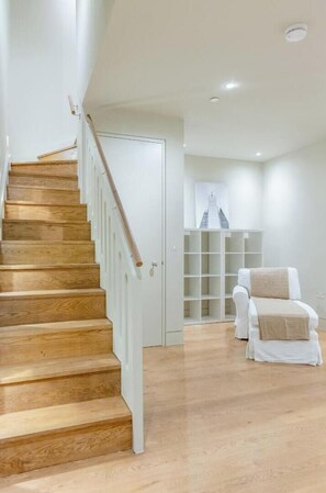 Eine Treppe mit Holzboden und weißen Geländern führt zu einem Raum mit einer weißen Tür, einem weißen Stuhl und einem weißen Regalschrank.