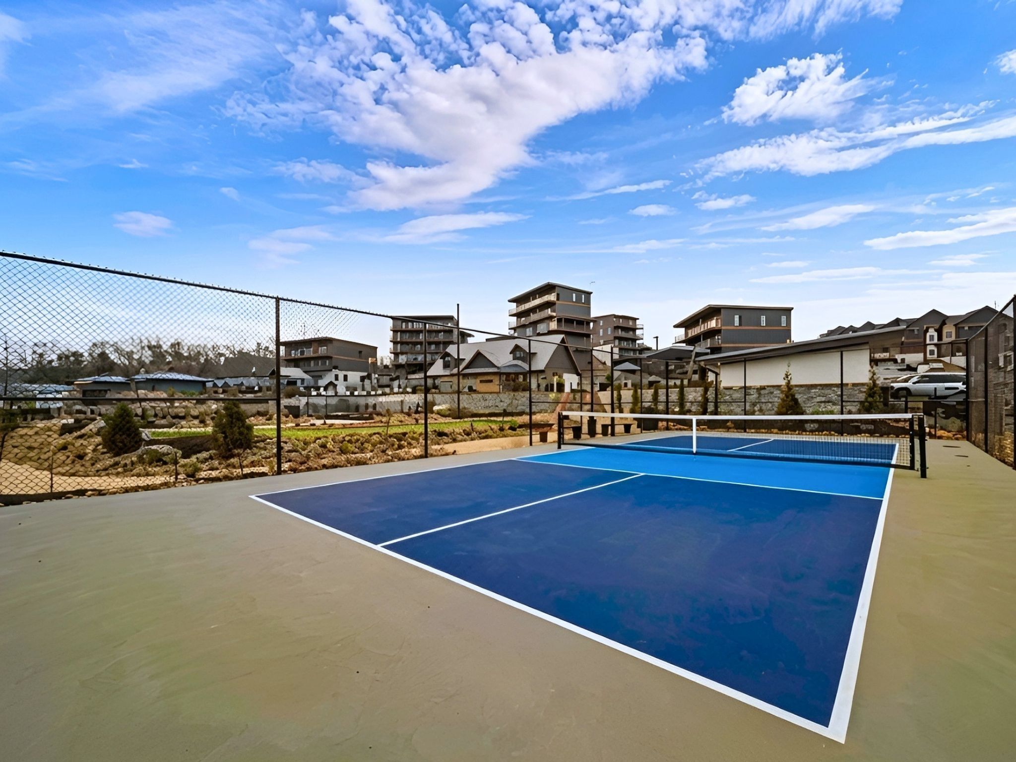 An outdoor tennis court with a chain-link fence, surrounded by residential buildings.