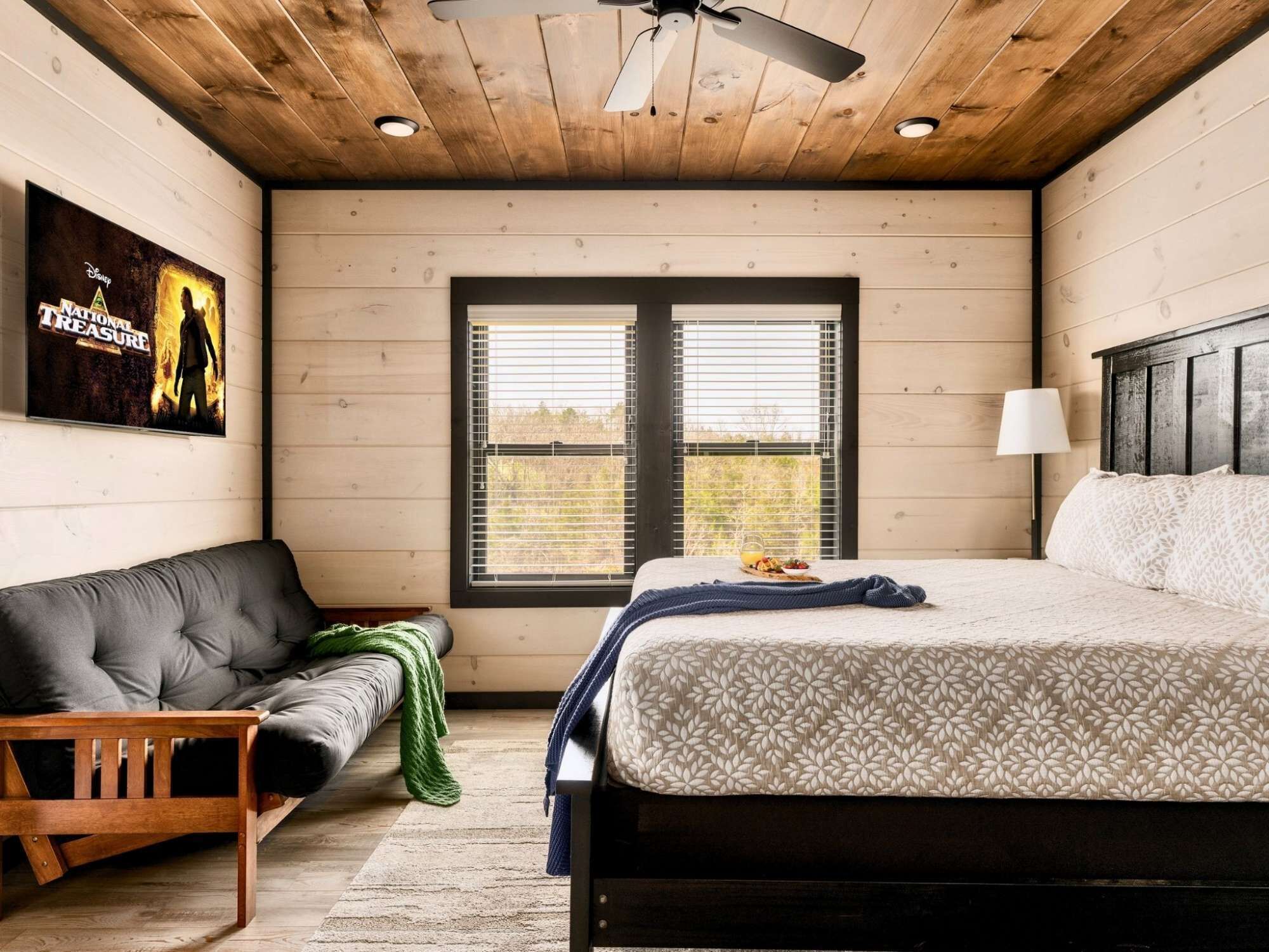 A bedroom with a bed, a couch, a window with blinds, a ceiling fan, and a poster on the wall.