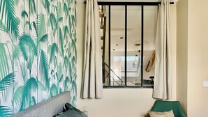 Una camera da letto con un letto, una poltrona verde, una finestra con tende e una parete decorata con un motivo a foglia.