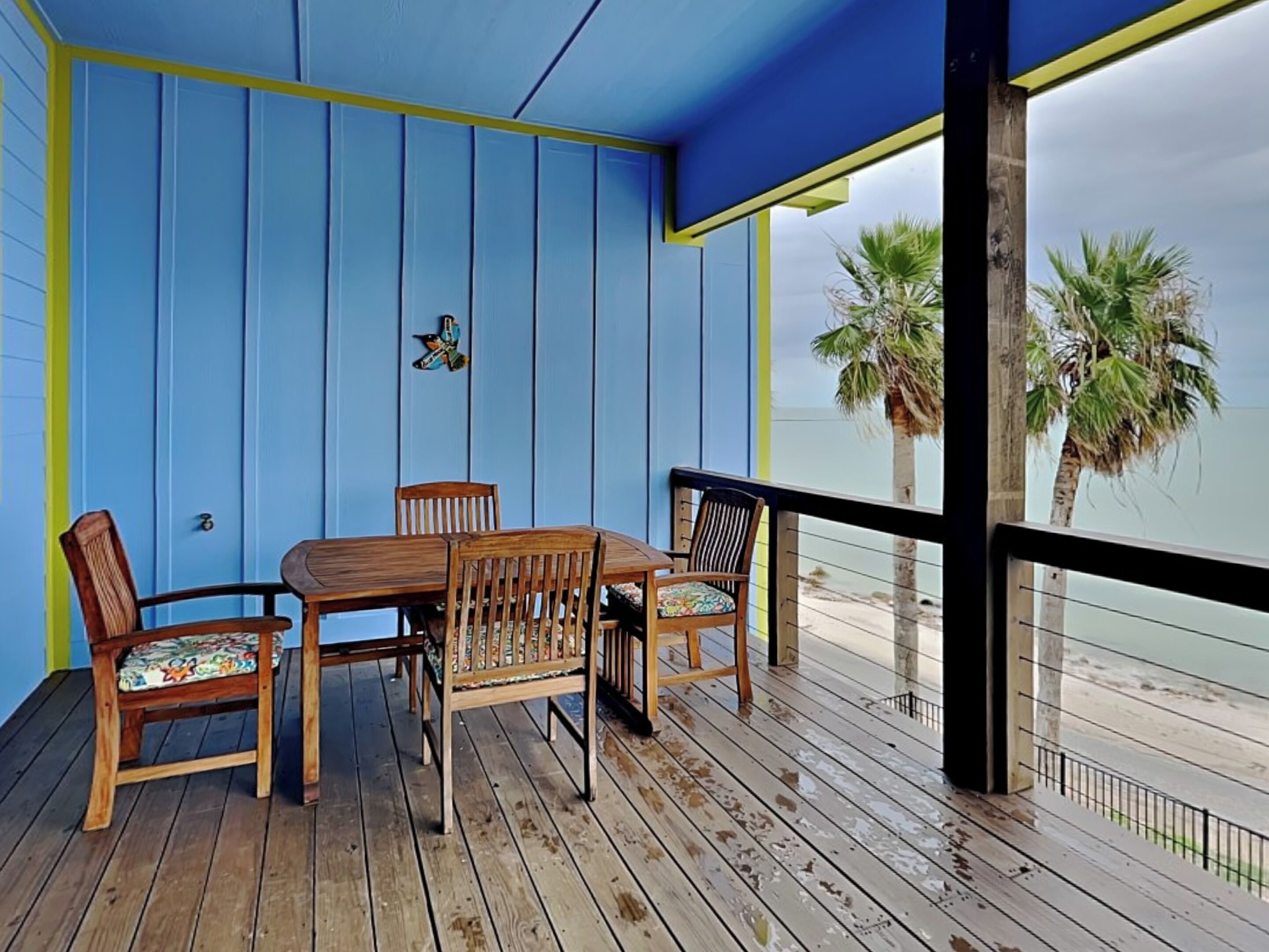 A wooden deck with a table and chairs, a blue wall with yellow trim, and a view of palm trees and water.