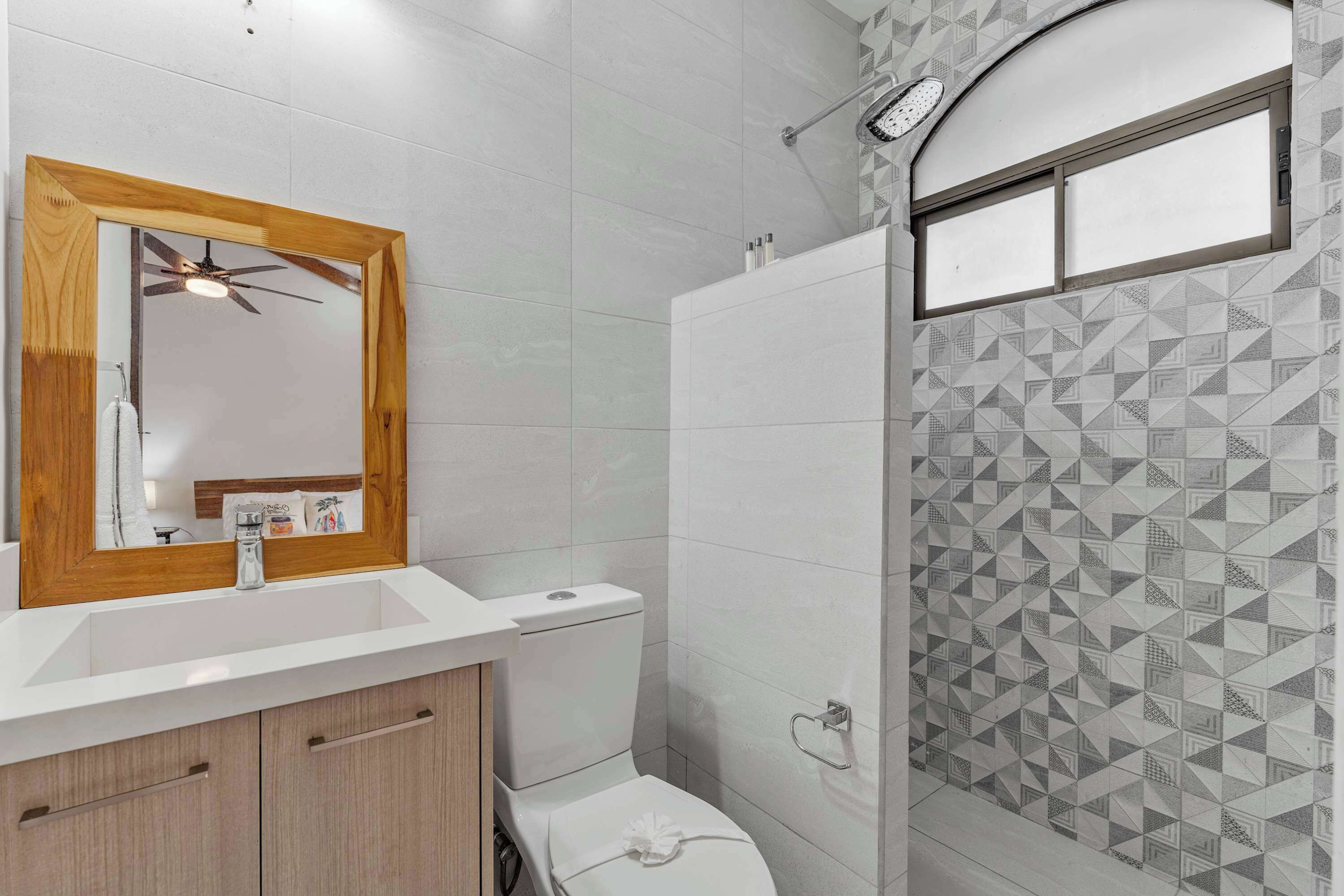 A bathroom with a wooden-framed mirror, a white sink, a toilet, and a shower with geometric-patterned tiles.