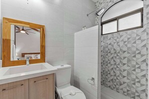 A bathroom with a wooden-framed mirror, a white sink, a toilet, and a shower with geometric-patterned tiles.