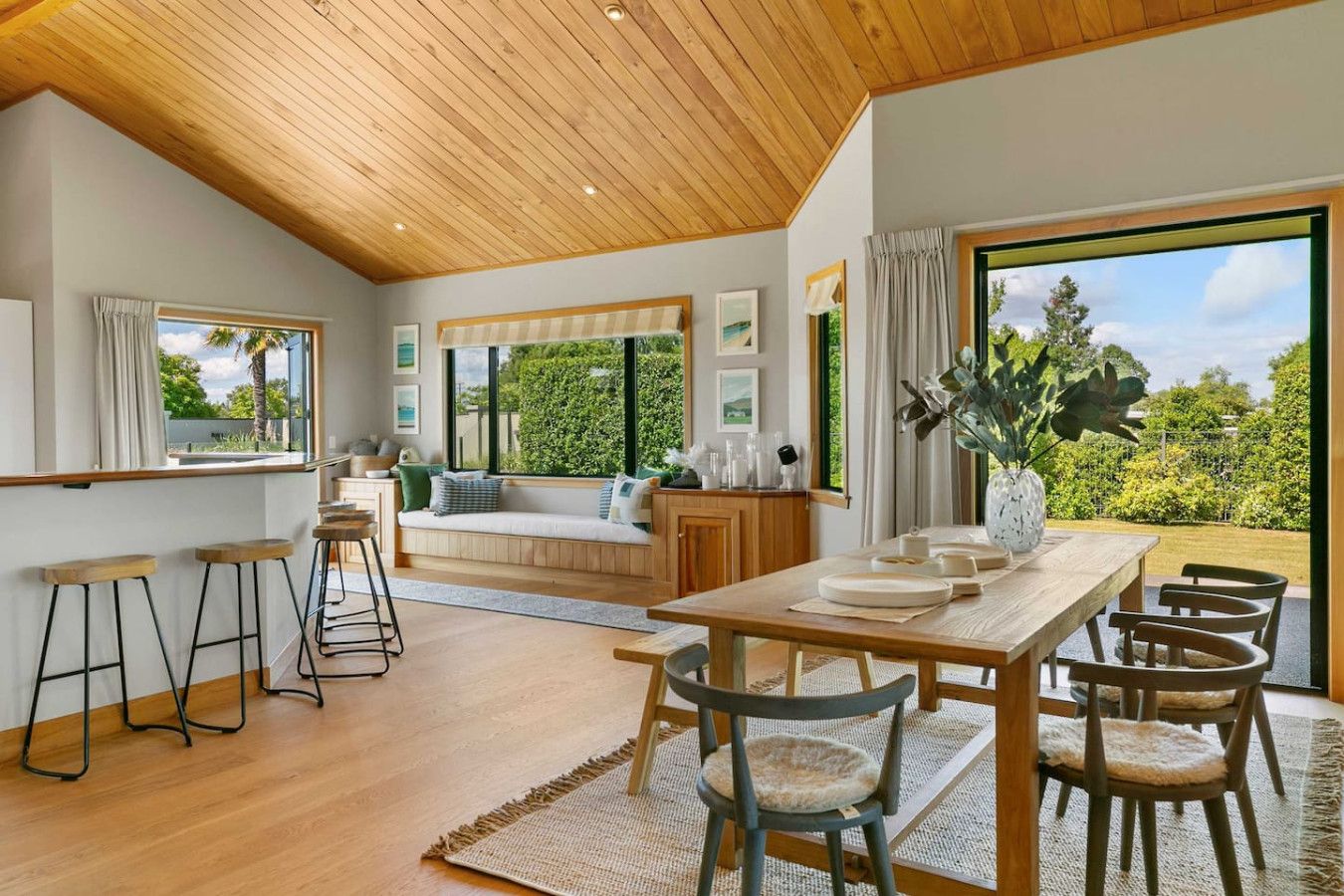 Amplia sala de estar con techo de madera, ventanas grandes que ofrecen vistas a la vegetación, una mesa de comedor con sillas y un rincón de descanso con sofá y cojines.