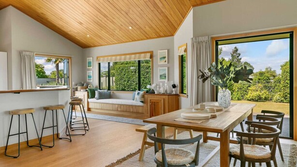 Un'ampia zona giorno con soffitto in legno, ampie finestre che offrono vista sul verde, un tavolo da pranzo con sedie e un angolo salotto con divano e cuscini.