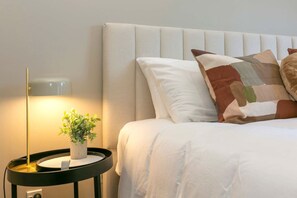 A neatly made bed with white bedding and a patchwork pillow, a bedside table with a lamp and a potted plant, and a wall-mounted light fixture.