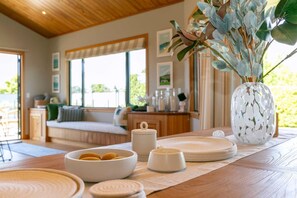 Un salon chaleureux avec un plafond en bois, une grande fenêtre et une vue sur la verdure. On y trouve un canapé, une table basse agrémentée d’objets décoratifs et un vase avec des plantes.