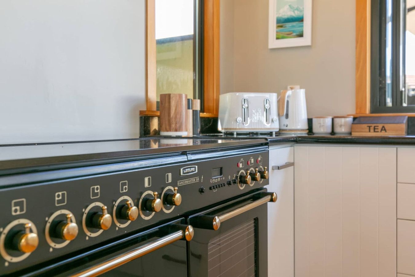 Een keuken met een zwarte kookplaat, een afzuigkap en een aanrecht met een broodrooster en een doosje met de tekst 'TEA'.
