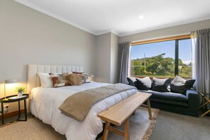 A bedroom with a large bed, a bench, a sofa, and a view of greenery through the window. - Watene Lodge - Spacious 5 Star Home with Pool (Taupo)