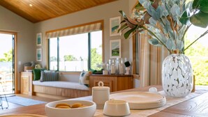 Un accogliente soggiorno con soffitto in legno, un'ampia finestra e vista sul verde circostante. Si nota un divano, un tavolino da caffè con oggetti decorativi e un vaso con piante.