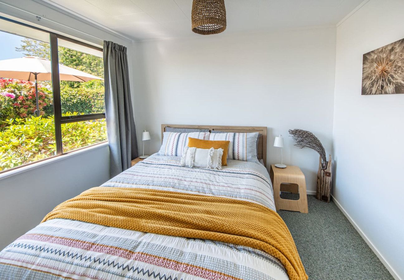 A bedroom with a large window, a bed with a patterned bedspread, a bedside table with a lamp, and a small wooden stool.