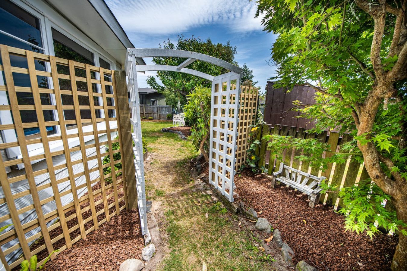 A garden with a wooden fence, a white trellis, and a tree.
