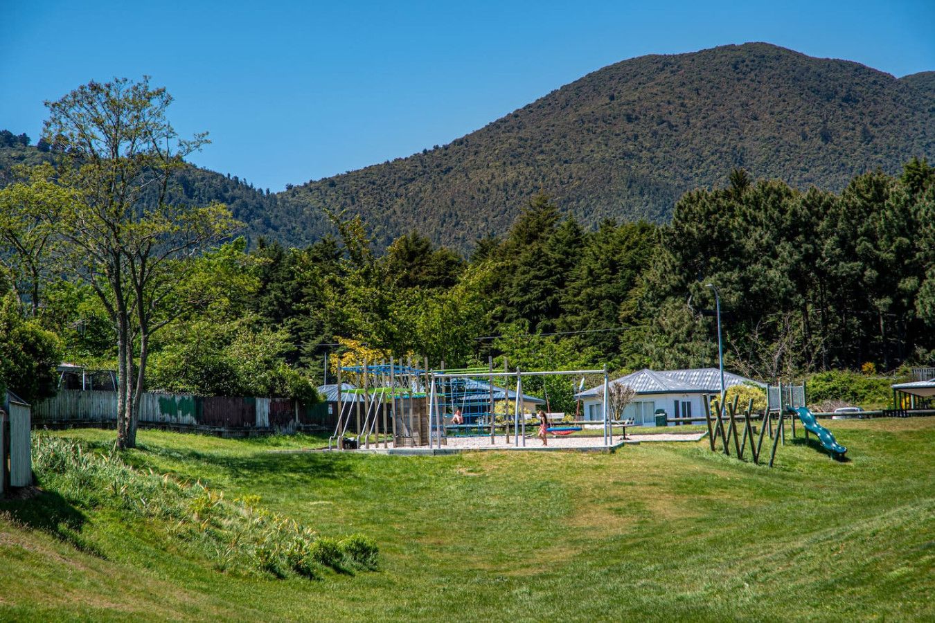 A playground with swings and a slide, surrounded by grass and trees, with a mountain in the background.