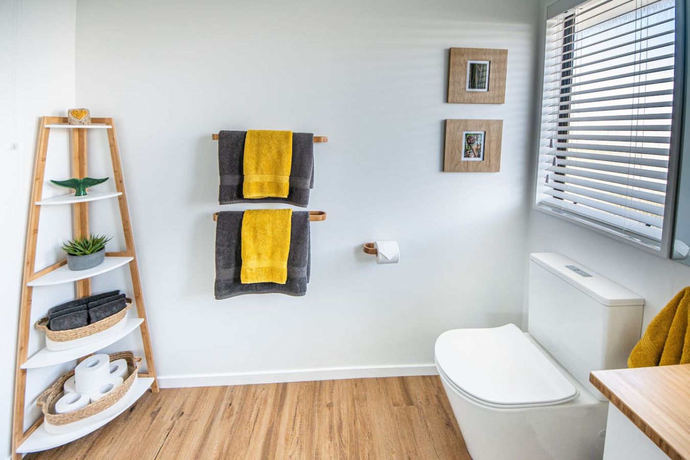 A bathroom with a wooden floor, white walls, a white toilet, a wooden shelf with towels and a plant, and two framed pictures on the wall.