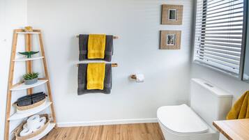 A bathroom with a wooden floor, white walls, a white toilet, a wooden shelf with towels and a plant, and two framed pictures on the wall.