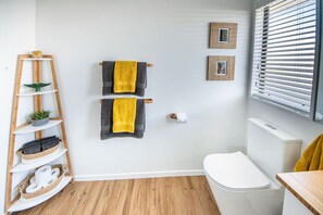 Une salle de bain avec un plancher en bois, des murs blancs, des toilettes blanches, une étagère en bois avec des serviettes et une plante, et deux tableaux encadrés accrochés au mur.
