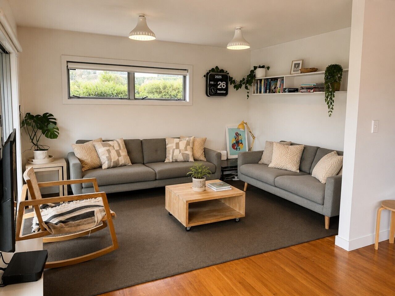 A living room with a grey sofa, a wooden coffee table, a rocking chair, and a bookshelf with plants.