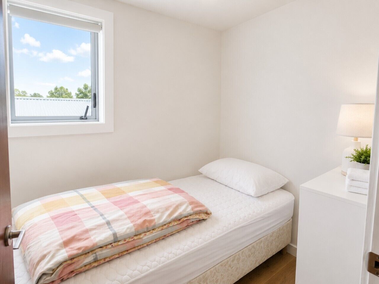 A single bed with a checkered blanket, a white pillow, a bedside table with a lamp, and a window with a view of the sky and trees.