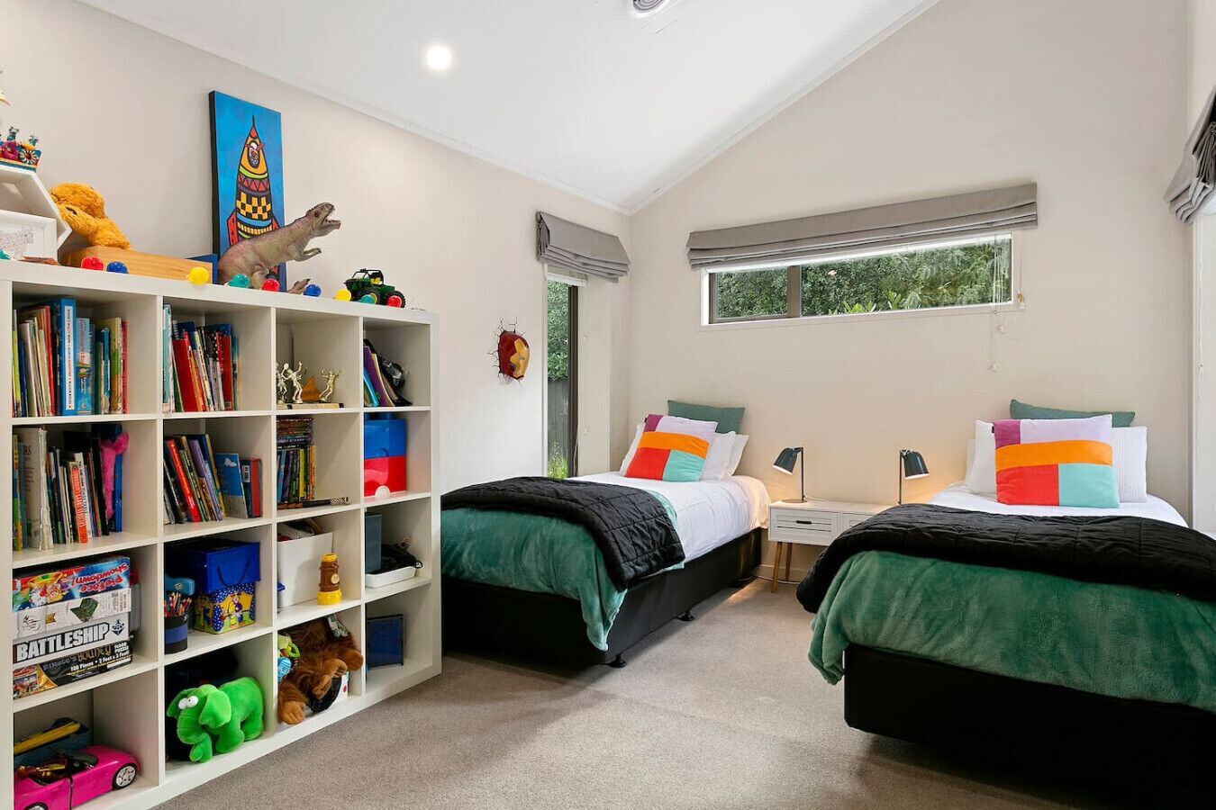 Une chambre avec deux lits, une bibliothèque remplie de livres et de jouets, et une fenêtre donnant sur un paysage verdoyant.