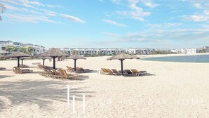 Una playa con tumbonas y sombrillas de paja, cielo despejado y un edificio de resort al fondo.