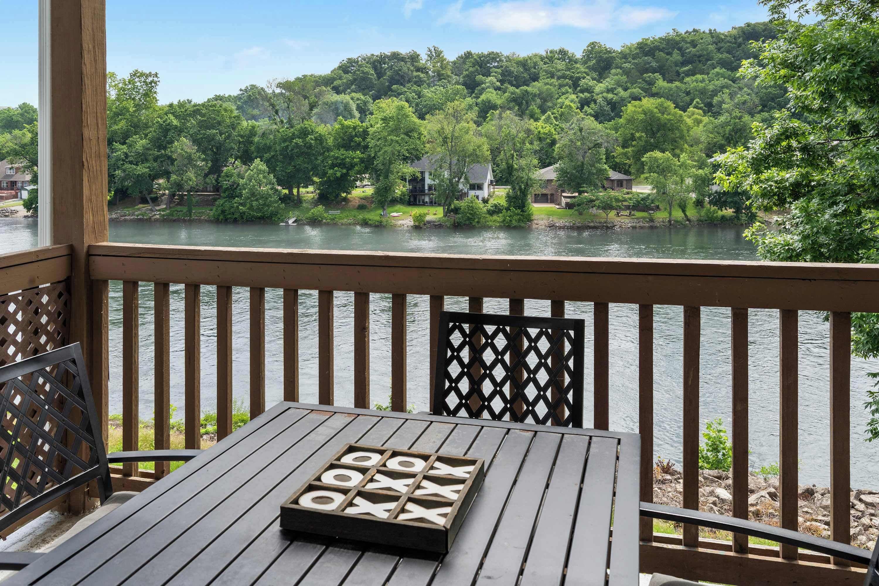 A wooden deck with a table and chairs, overlooking a river with houses and greenery in the background.