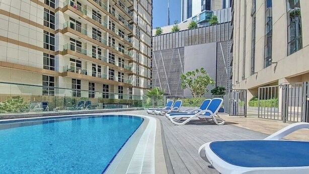 A swimming pool with lounge chairs, a wooden deck, and a high-rise building in the background. - Livbnb-2B+Maid's in Dunya, 5Min Walk to Dubai Mall (Dubai)