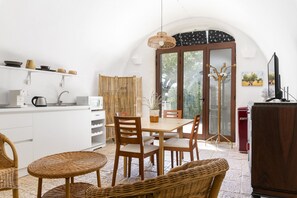 Cozinha com armários brancos, área de refeições com cadeiras de madeira e uma mesa, uma cadeira de vime, uma televisão e um eletrodoméstico vermelho.