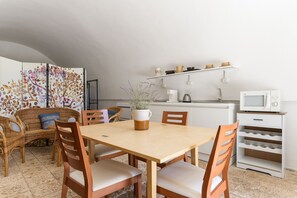A dining area with a wooden table and chairs, a wicker sofa, a white cabinet with shelves, and a microwave.