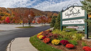 Señal de 'Sugarbush BeBetterHere' en Lincoln Peak Village, con vistas a árboles de colores otoñales y un estacionamiento al fondo.