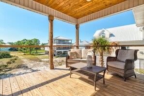 A covered outdoor area with a wooden deck, wicker furniture, and a view of a residential area with houses and a clear sky. - Sunset Blessing - 207 Arias Ct 4 Bedroom Home (Dauphin Island)