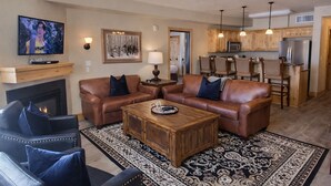 A living room with leather sofas, a coffee table, and a fireplace. There is a kitchen area with wooden cabinets and a refrigerator.