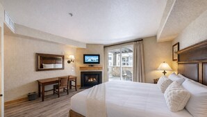 A hotel room with a large bed, a desk, a fireplace, a television, and a balcony with a view.