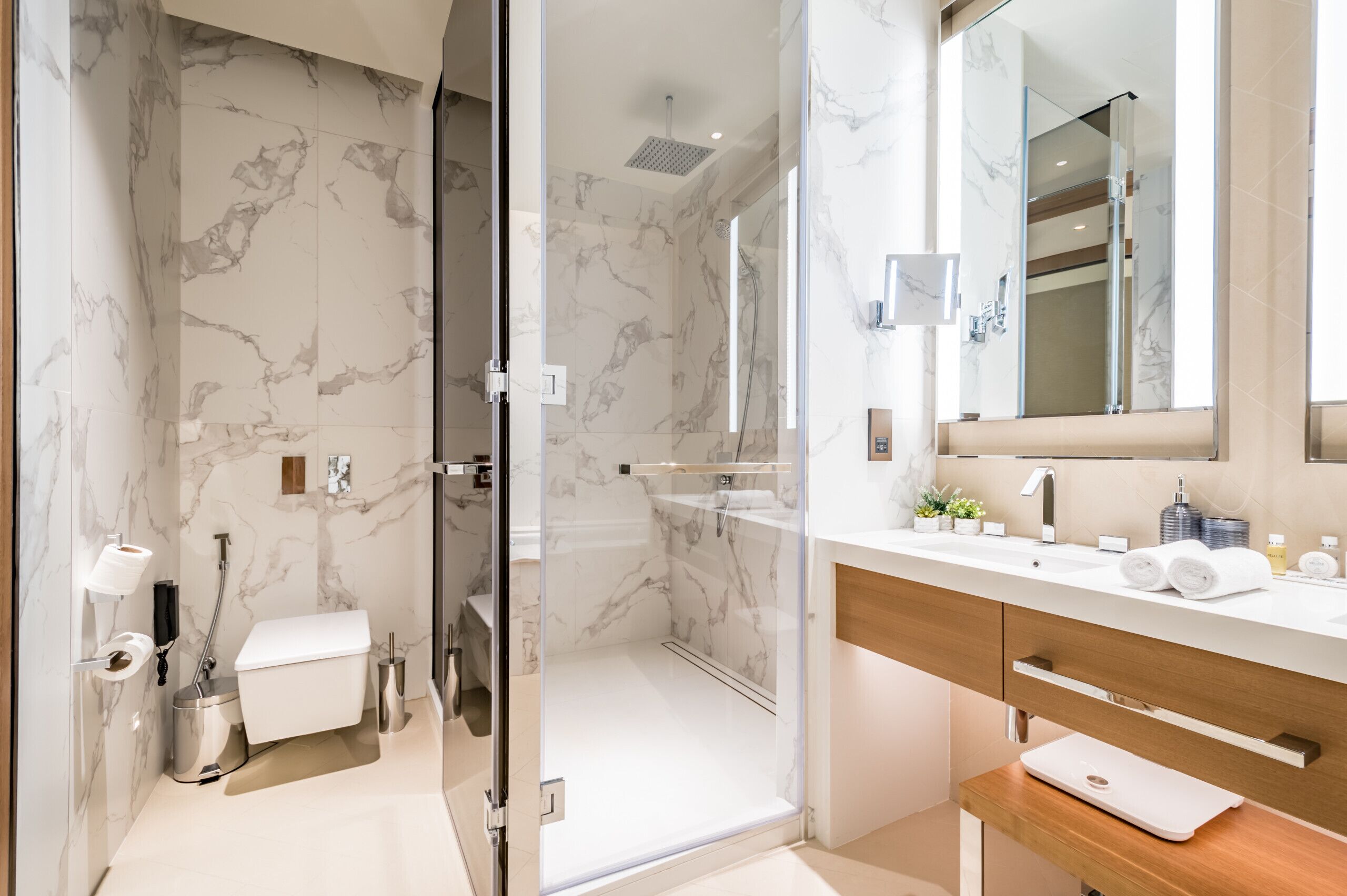 A modern bathroom with marble walls, a glass-enclosed shower, a wooden vanity, and a large mirror.