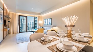 A modern hotel room with a dining area set for four, large windows with a city view, a sofa, and a lamp.