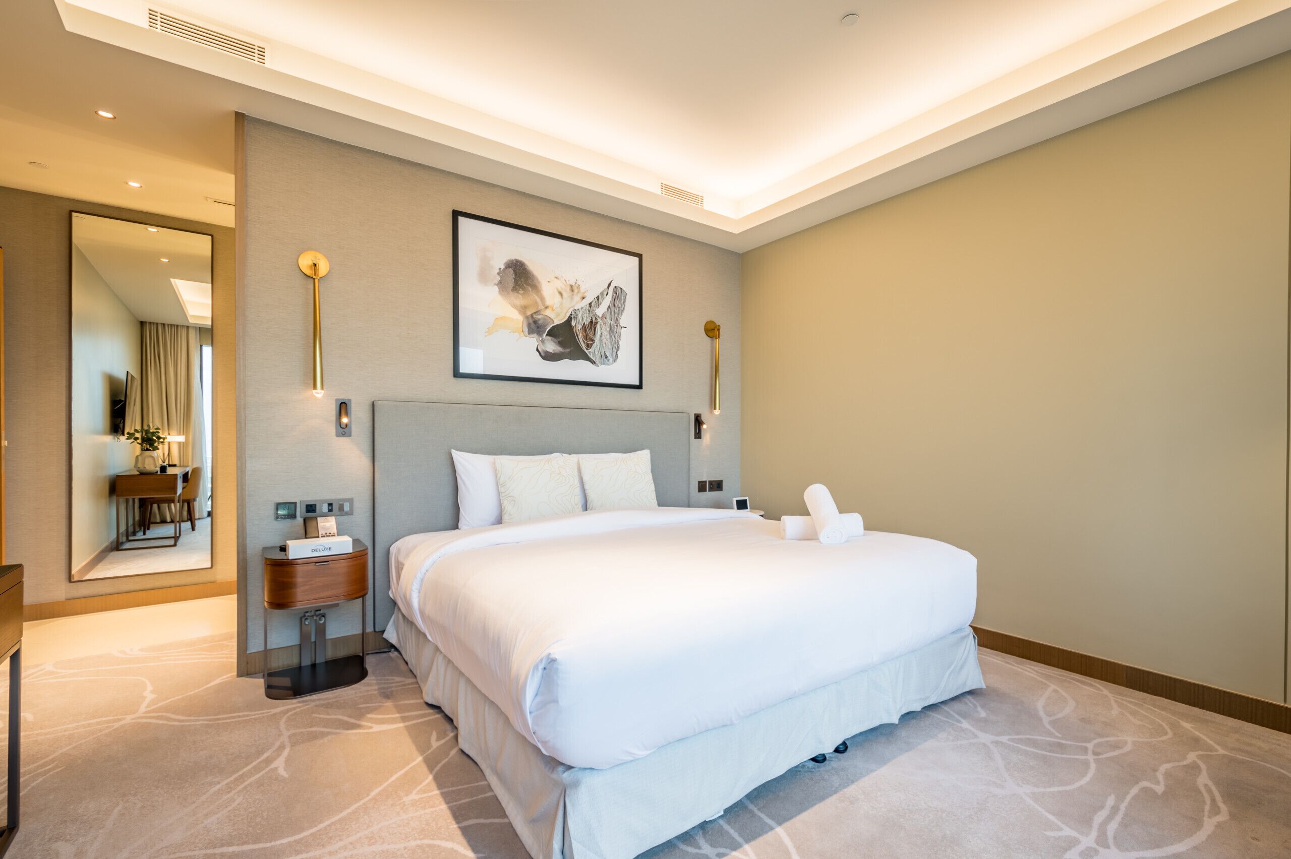 Une chambre d’hôtel moderne avec un grand lit, des tables de chevet, un miroir et une œuvre d’art encadrée accrochée au mur.