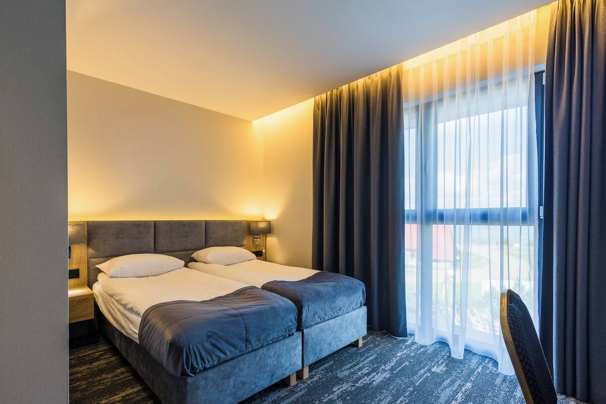 A hotel room with a double bed, a bedside table, a chair, and a large window with curtains.