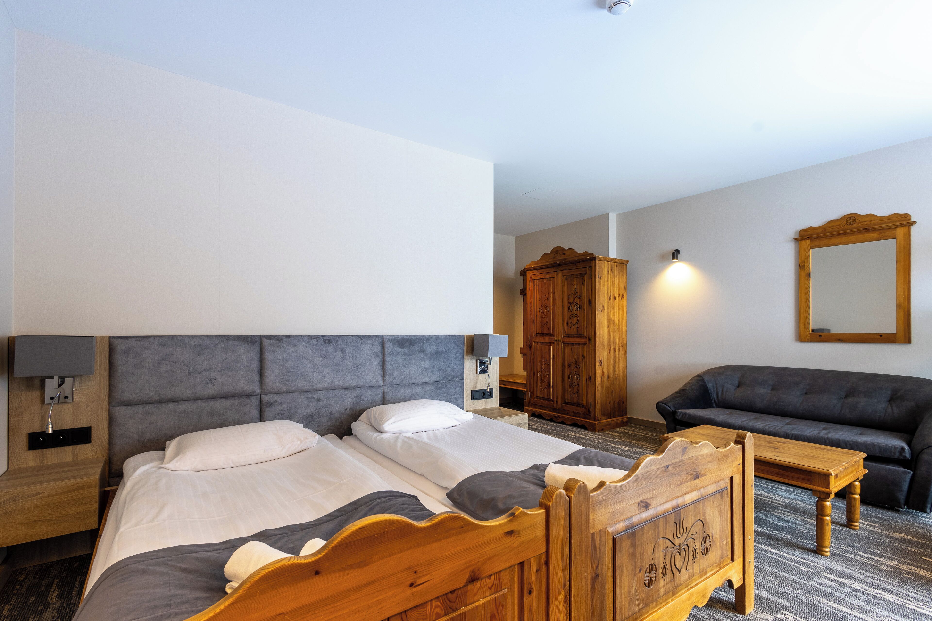 Une chambre d’hôtel avec un lit en bois, un canapé gris, une table de chevet en bois, une armoire en bois et un miroir au mur.