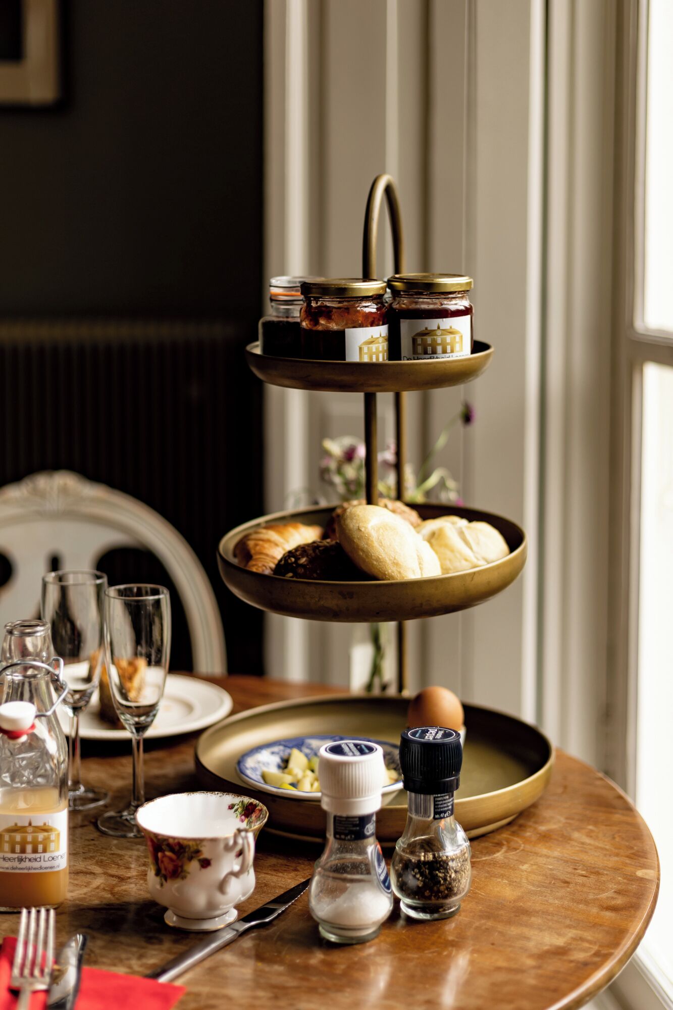 A tiered serving tray with pastries and condiments on a table, accompanied by glasses and a teapot.