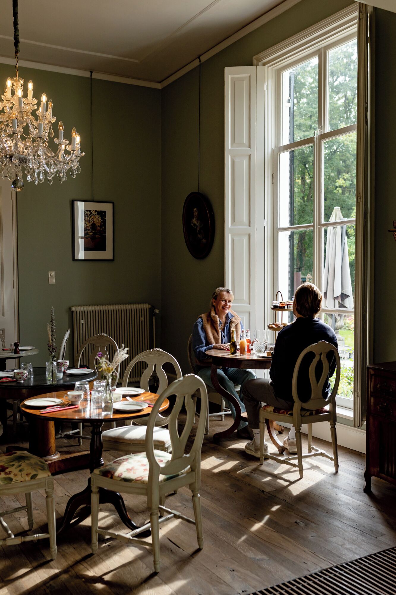 A dining room with a chandelier, a table set for two, and a view of the outdoors through a large window.