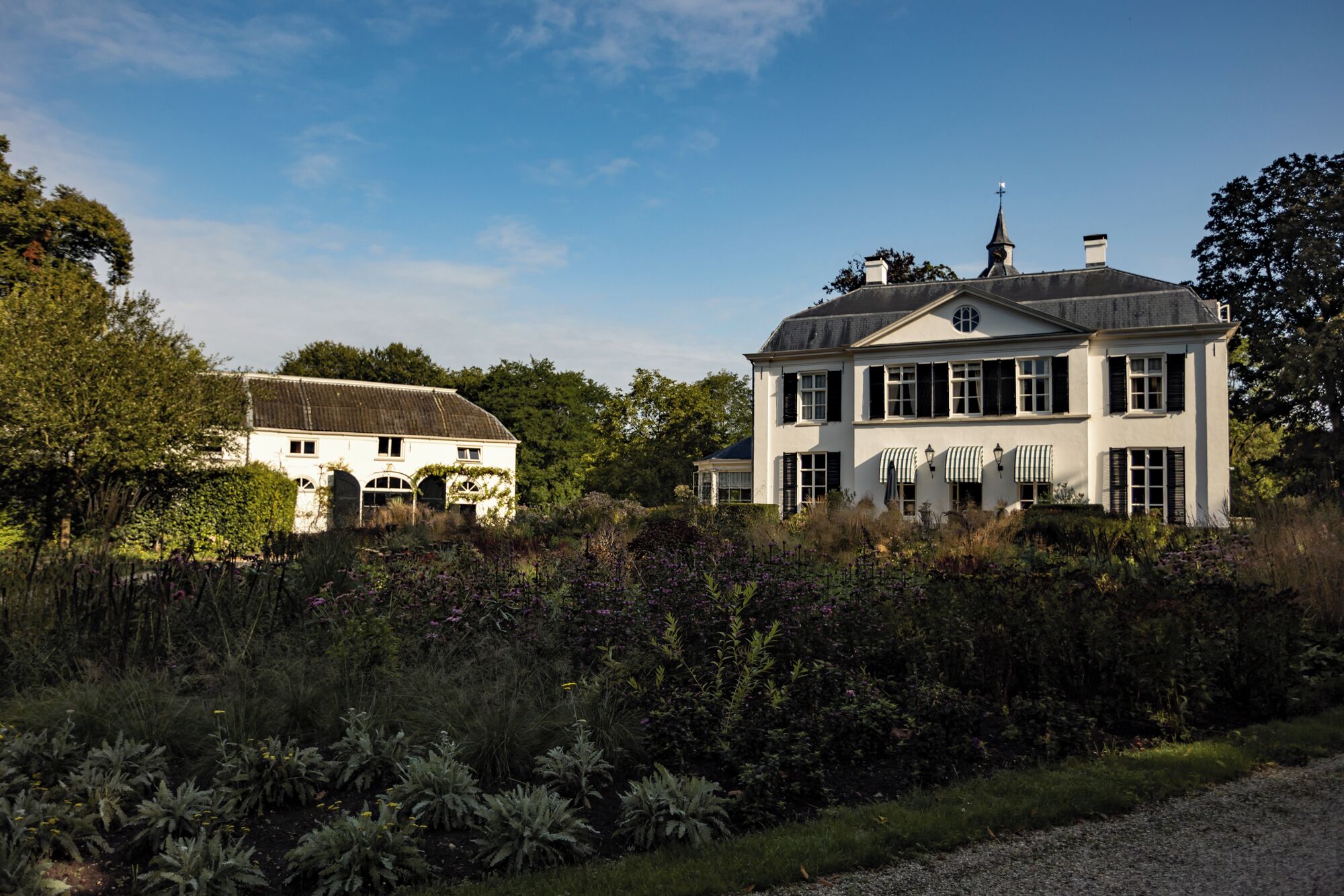 A large white building with multiple windows and a chimney, surrounded by lush greenery and a gravel path.